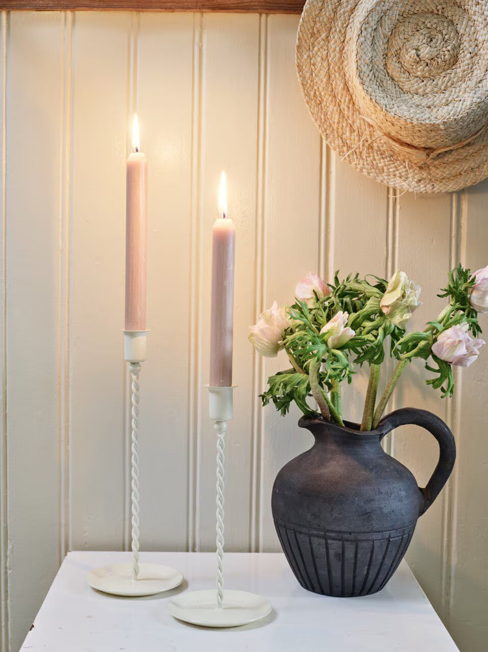 witte metalen kandelaar van Strömshaga met gedraaide steel op een witte tafel naast een donkere brocante kruik met bloemen.