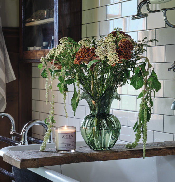Groene glazen vaas met veldboeket op houten badplank in een vintage badkamer met vrijstaand bad en witte tegels