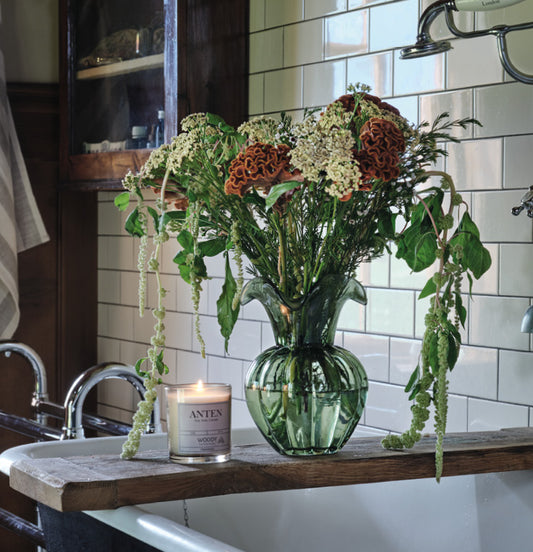 Groene glazen vaas met veldboeket op houten badplank in een vintage badkamer met vrijstaand bad en witte tegels