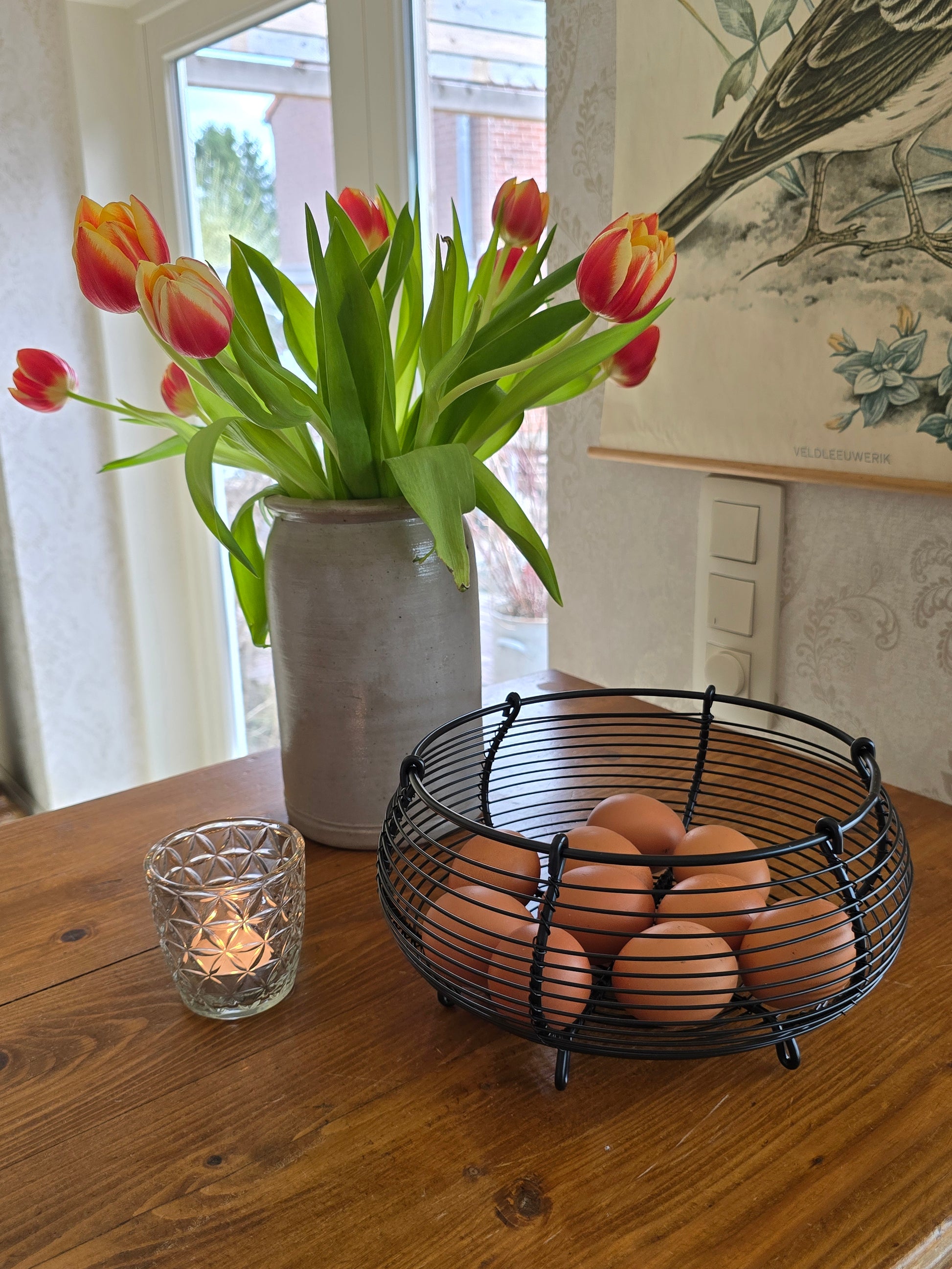 Zwart metalen eiermandje met bruine eieren op houten keukentoog naast tulpen in vaas
