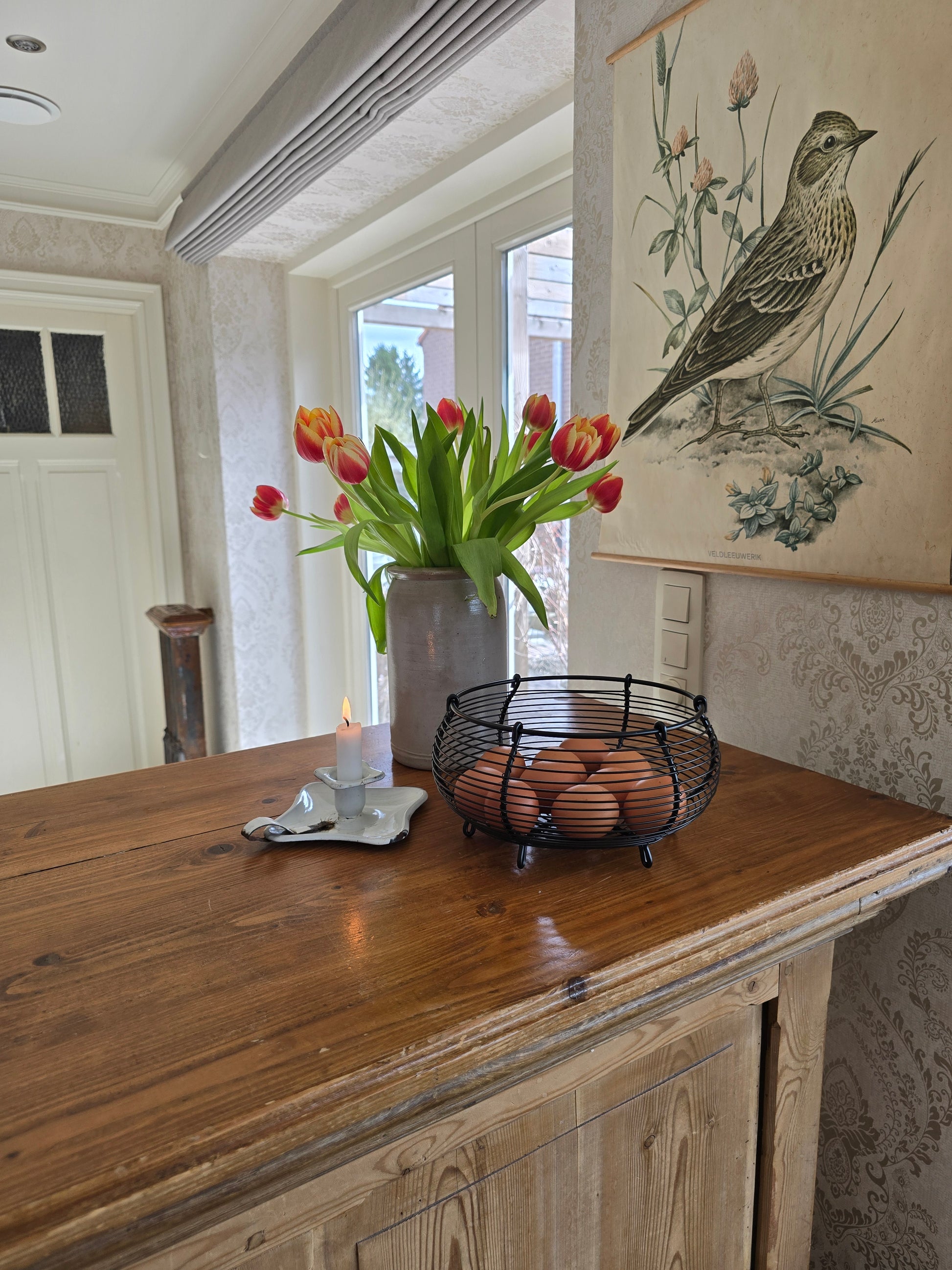 Zwart metalen eiermandje met bruine eieren op houten keukentoog naast tulpen in vaas en brocante kaarsenhouder